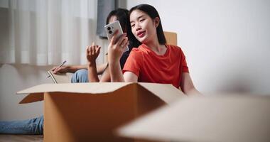 Handheld low angle view shot, Two young Asian women sit with backs to each other on the floor and discuss ideas for designing a room together in their new home. Moving to a new home concept video