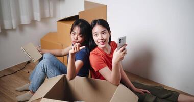 Handheld high angle view shot, Two young Asian women sit with backs to each other on the floor and discuss ideas for designing a room together in their new home. Moving to a new home concept video