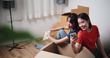 Handheld high angle view shot, Two young Asian women sit with backs to each other on the floor and discuss ideas for designing a room together in their new home. Moving to a new home concept video