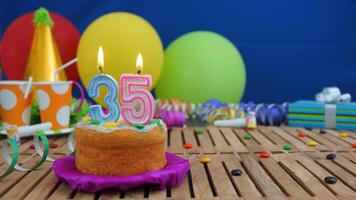 Birthday cake 35 with candles on rustic wooden table with background of colorful balloons, gifts, plastic cups and candies with blue wall in the background. Focus is on cake photo