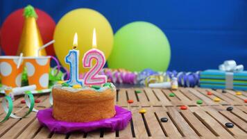 Birthday cake 12 with candles on rustic wooden table with background of colorful balloons, gifts, plastic cups and candies with blue wall in the background. Focus is on cake photo