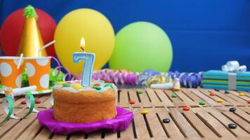 Birthday cake 7 with candles on rustic wooden table with background of colorful balloons, gifts, plastic cups and candies with blue wall in the background. Focus is on cake photo