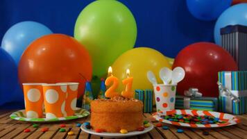 Birthday cake 21 on rustic wooden table with background of colorful balloons, gifts, plastic cups and plastic plate with candies and blue wall in the background photo