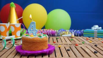 Birthday cake 4 with candles on rustic wooden table with background of colorful balloons, gifts, plastic cups and candies with blue wall in the background. Focus is on cake photo