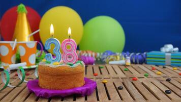 Birthday cake 38 with candles on rustic wooden table with background of colorful balloons, gifts, plastic cups and candies with blue wall in the background. Focus is on cake photo