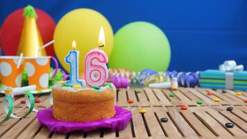 Birthday cake 16 with candles on rustic wooden table with background of colorful balloons, gifts, plastic cups and candies with blue wall in the background. Focus is on cake photo