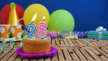 Birthday cake 26 with candles on rustic wooden table with background of colorful balloons, gifts, plastic cups and candies with blue wall in the background. Focus is on cake photo