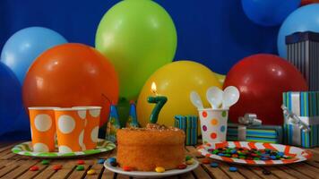 Birthday cake 7 on rustic wooden table with background of colorful balloons, gifts, plastic cups and plastic plate with candies and blue wall in the background photo