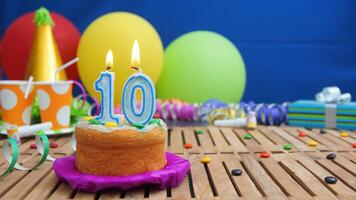Birthday cake 10 with candles on rustic wooden table with background of colorful balloons, gifts, plastic cups and candies with blue wall in the background. Focus is on cake photo