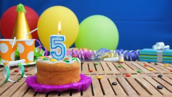 Birthday cake 5 with candles on rustic wooden table with background of colorful balloons, gifts, plastic cups and candies with blue wall in the background. Focus is on cake photo