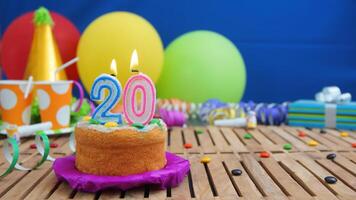 Birthday cake 20 with candles on rustic wooden table with background of colorful balloons, gifts, plastic cups and candies with blue wall in the background. Focus is on cake photo