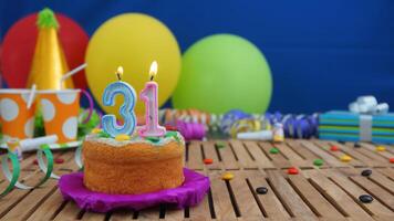Birthday cake 31 with candles on rustic wooden table with background of colorful balloons, gifts, plastic cups and candies with blue wall in the background. Focus is on cake photo