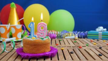 Birthday cake 19 with candles on rustic wooden table with background of colorful balloons, gifts, plastic cups and candies with blue wall in the background. Focus is on cake photo