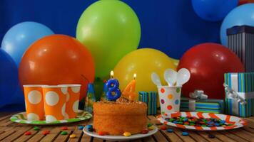 Birthday cake 34 on rustic wooden table with background of colorful balloons, gifts, plastic cups and plastic plate with candies and blue wall in the background photo