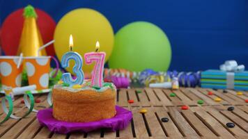 Birthday cake 37 with candles on rustic wooden table with background of colorful balloons, gifts, plastic cups and candies with blue wall in the background. Focus is on cake photo