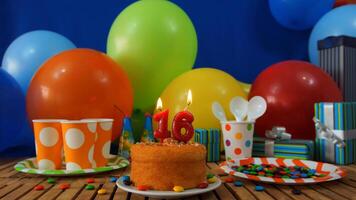 Birthday cake 16 on rustic wooden table with background of colorful balloons, gifts, plastic cups and plastic plate with candies and blue wall in the background photo