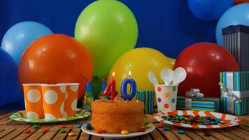 Birthday cake 40 on rustic wooden table with background of colorful balloons, gifts, plastic cups and plastic plate with candies and blue wall in the background photo