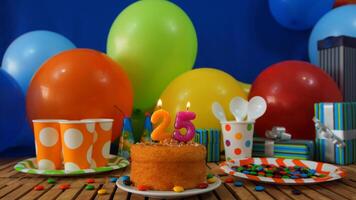 Birthday cake 25 on rustic wooden table with background of colorful balloons, gifts, plastic cups and plastic plate with candies and blue wall in the background photo