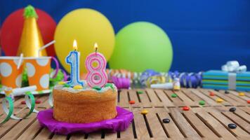 Birthday cake 18 with candles on rustic wooden table with background of colorful balloons, gifts, plastic cups and candies with blue wall in the background. Focus is on cake photo