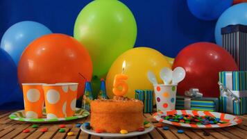 Birthday cake 5 on rustic wooden table with background of colorful balloons, gifts, plastic cups and plastic plate with candies and blue wall in the background photo