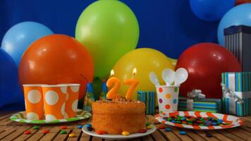 Birthday cake 27 on rustic wooden table with background of colorful balloons, gifts, plastic cups and plastic plate with candies and blue wall in the background photo