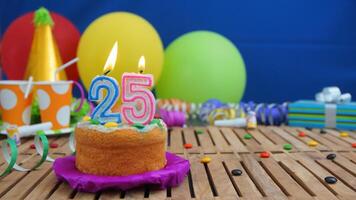 Birthday cake 25 with candles on rustic wooden table with background of colorful balloons, gifts, plastic cups and candies with blue wall in the background. Focus is on cake photo