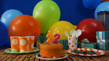 Birthday cake 22 on rustic wooden table with background of colorful balloons, gifts, plastic cups and plastic plate with candies and blue wall in the background photo