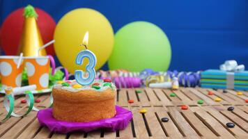 Birthday cake 3 with candles on rustic wooden table with background of colorful balloons, gifts, plastic cups and candies with blue wall in the background. Focus is on cake photo