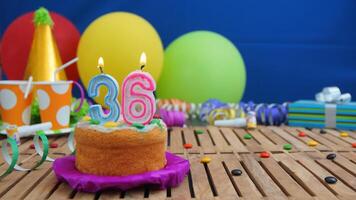 Birthday cake 36 with candles on rustic wooden table with background of colorful balloons, gifts, plastic cups and candies with blue wall in the background. Focus is on cake photo