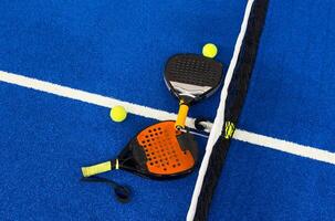 A padel or paddle tennis racket and a bright yellow tennis ball lie on a blue court. The net and a blurred figure of a player are visible in the background. photo