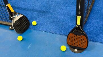 A padel or paddle tennis racket and a bright yellow tennis ball lie on a blue court. The net and a blurred figure of a player are visible in the background. photo