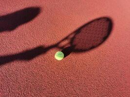 High angle of yellow ball placed on court and covered with shadow of crop person holding padel racket photo