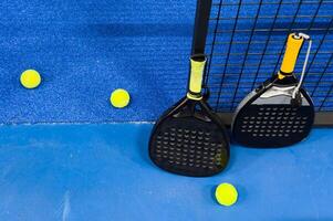 A padel or paddle tennis racket and a bright yellow tennis ball lie on a blue court. The net and a blurred figure of a player are visible in the background. photo