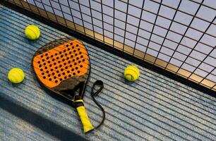 A padel or paddle tennis racket and a bright yellow tennis ball lie on a blue court. The net and a blurred figure of a player are visible in the background. photo