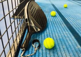 Padel racket and yellow ball behind net on a green court grass turf outdoors at sunset. Paddle is a racquet game. Professional sport concept with space for text photo