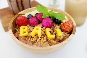 homemade granola with yogurt and fresh watermelon with pineapple carved for Bali word, dragon fruit and strawberry topping in wooden bowl con white table in cafe photo