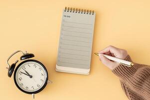 Creative layout made with woman's hand holding white pen, To do list in spiral notepad and retro alarm clock. Trendy pastel background, flat lay style. Planning concept. photo