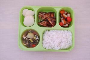 A green tray with five compartments containing white rice, curry, sliced cucumbers and tomatoes, a boiled egg, quail eggs, fried chicken, and pieces of meat photo