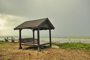 A wooden gazebo by a calm beach surrounded the sea beneath cloudy skies that creates a peaceful setting that embodies simplicity and solitude in the coastal landscape. photo
