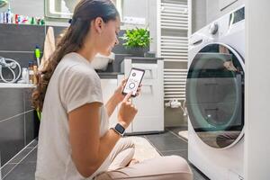 Young woman using mobile phone control via wi-fi and app for washing machine in home. Homesmart and new technology concept. High quality photo