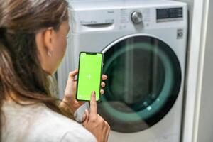 Young woman using mobile phone control via wi-fi and app for washing machine in home. Homesmart app, chroma key green screen. High quality photo