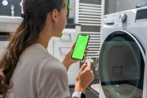 Young woman using mobile phone control via wi-fi and app for washing machine in home. Homesmart app, chroma key green screen. High quality photo