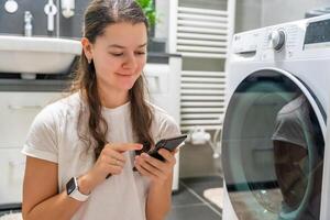 Young woman using mobile phone control via wi-fi and app for washing machine in home. Homesmart and new technology concept. High quality photo