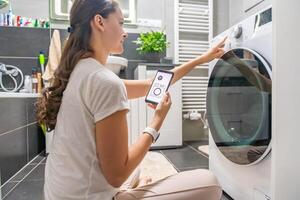 Young woman using mobile phone control via wi-fi and app for washing machine in home. Homesmart and new technology concept. High quality photo