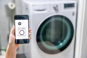 Young woman using mobile phone control via wi-fi and app for washing machine in home. Homesmart and new technology concept. High quality photo
