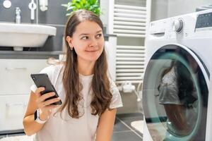 Young woman using mobile phone control via wi-fi and app for washing machine in home. Homesmart and new technology concept. High quality photo