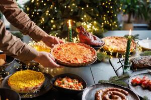 Man places Christmas quiche pie on festive table surrounded by many dishes in cozy evening atmosphere with Christmas tree and garlands photo