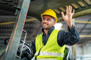 man in a yellow vest and a hard hat is waving from the cab of a forklift photo