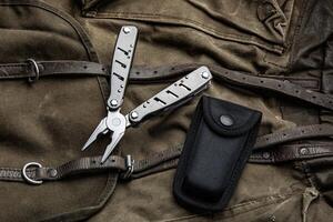 A multi-tool with pliers is placed on a brown canvas bag with leather straps, accompanied by a black nylon sheath, showcasing its utility and rugged design photo