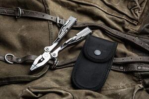 A multi-tool with pliers is placed on a brown canvas bag with leather straps, accompanied by a black nylon sheath, showcasing its utility and rugged design photo
